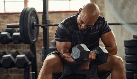 Muscular guy in sportswear lifting dumbbell while sitting on bench at crossfit gym. Mature african american athlete using dumbbell during a workout. Strong man under physical exertion pumping up bicep muscule with heavy weight.